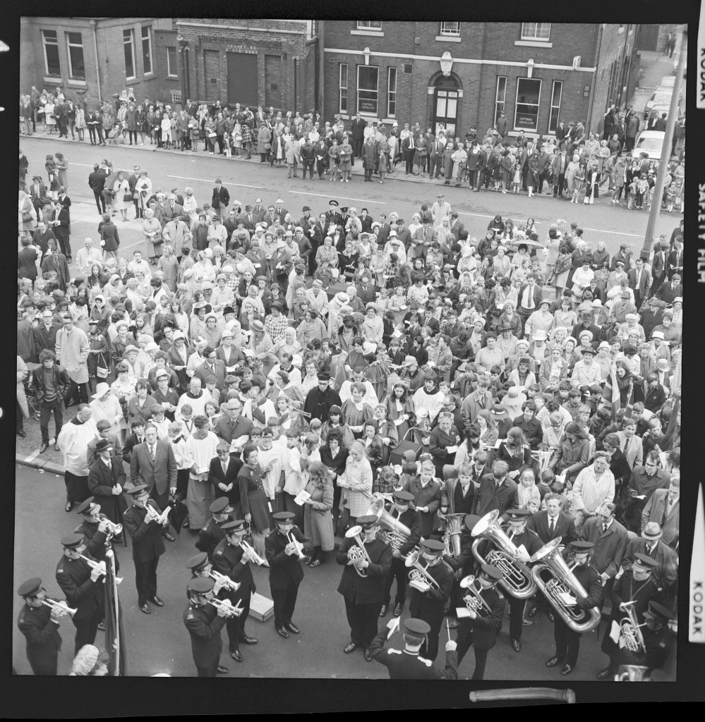 A crowd contaiing clergymembers, men, women and children on a high street with a road dividing the crowd watching a brass band